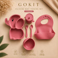 A 5-piece pink silicone baby feeding set by GOKIT, including an elephant-shaped divided plate, a small cup, a spoon, a fork, and a bib, arranged on a beige background with decorative dried flowers and a gold ribbon.