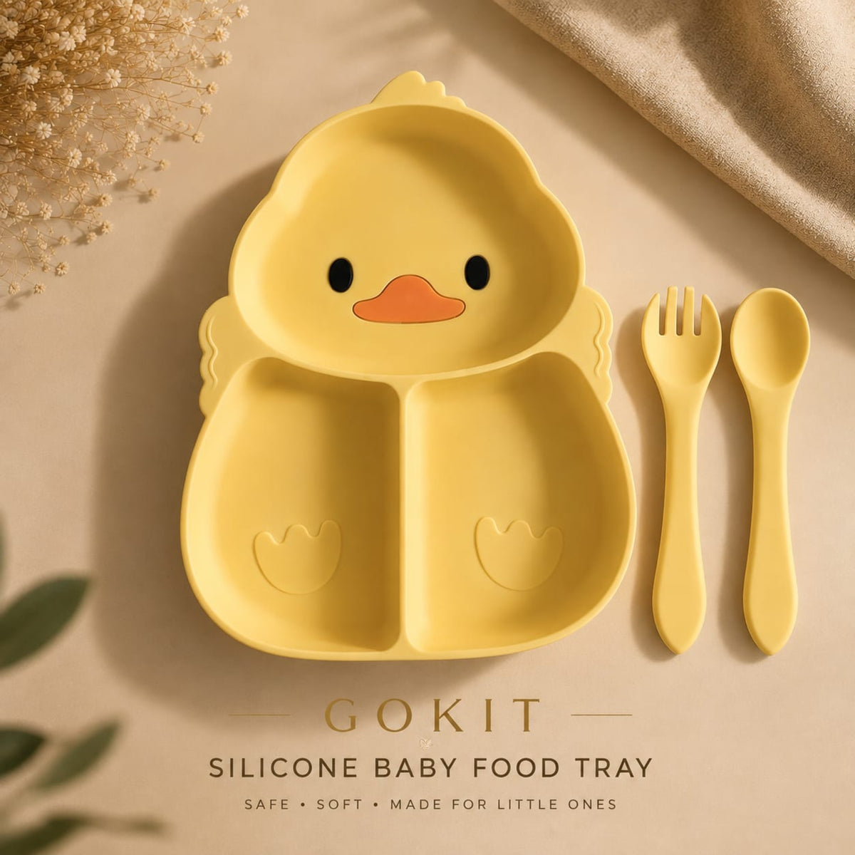 Yellow silicone baby food tray shaped like a duck face, shown with matching yellow fork and spoon on a beige surface with decorative dried flowers and fabric. Text at the bottom reads 'GOKIT SILICONE BABY FOOD TRAY'.