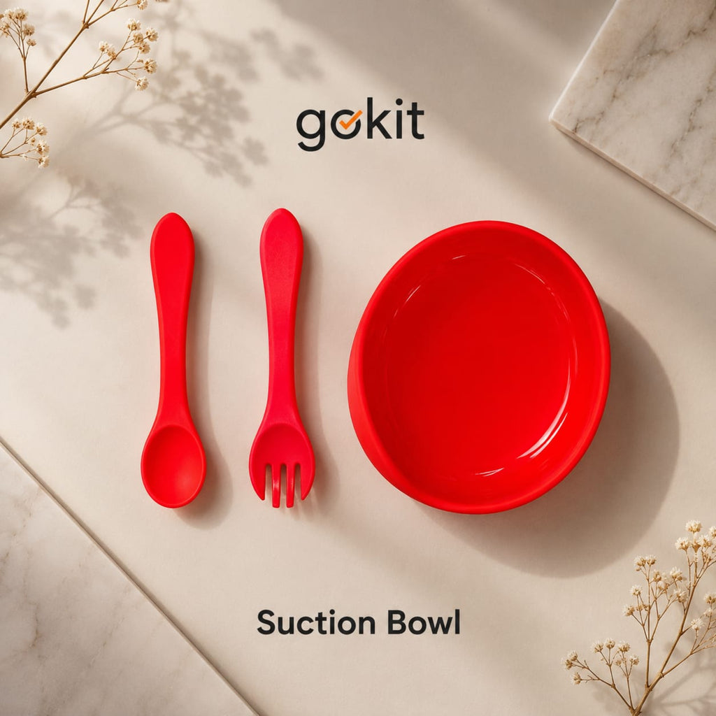 A red silicone suction bowl set with a spoon and fork, arranged on a light beige marble surface with dried flowers. The brand name 'gokit' is visible at the top.